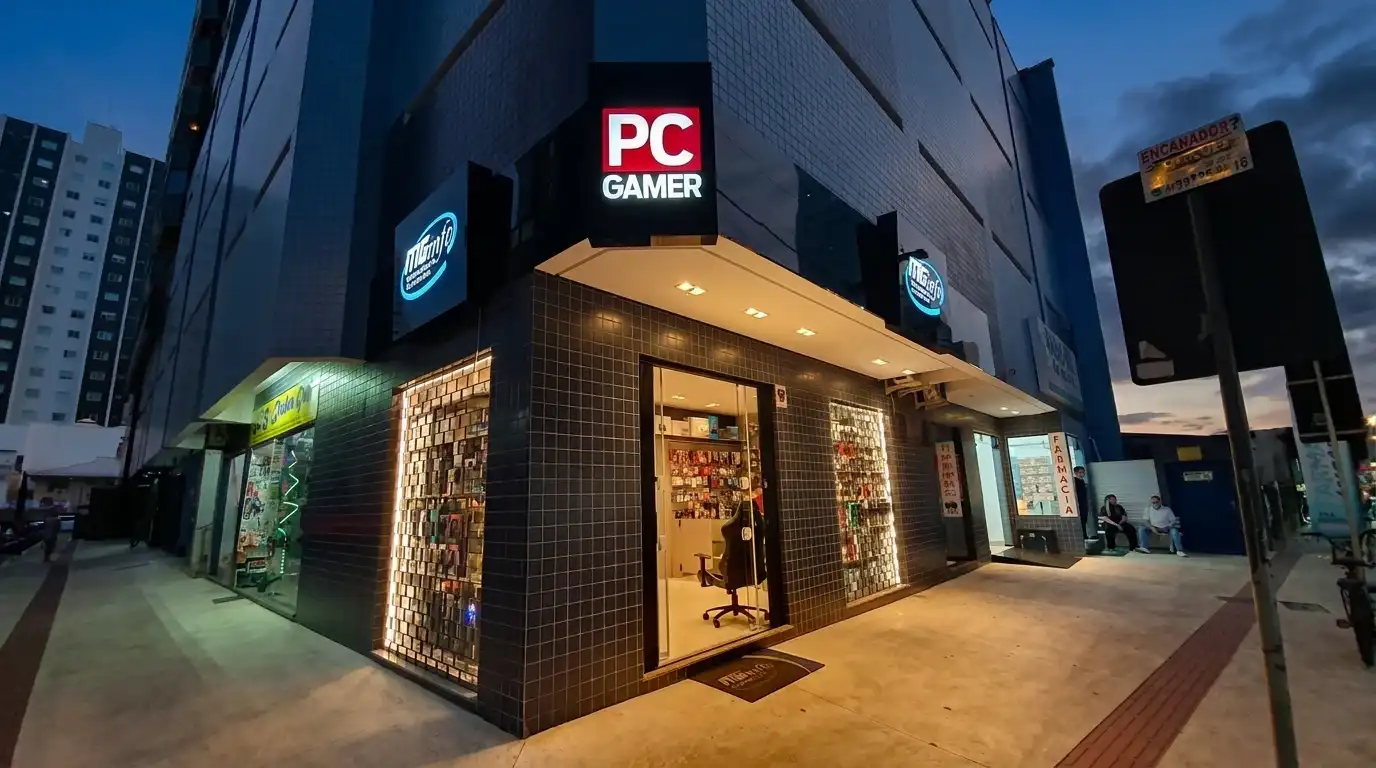 A esquina da MGinfo ao anoitecer: fachada de pastilha escura, a placa vermelha PC GAMER no alto, os letreiros redondos azuis e as vitrines iluminadas